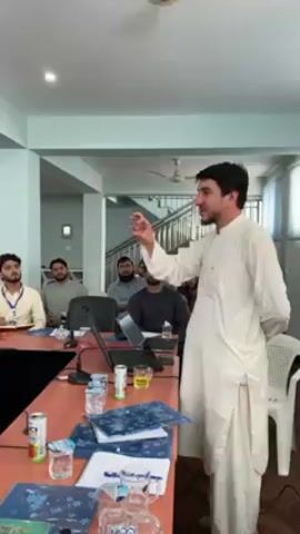 In an indoor setting, likely a conference or training room in Shurandam, Afghanistan, a man in a light-colored shalwar kameez stands and addresses a group of men. He is positioned to the right of a long, reddish-brown wooden table, actively gesturing with his right hand as he speaks. His audience, consisting of several men, is seated around the table, attentively listening. Some audience members are also wearing traditional attire, while others are in casual shirts.

The table is furnished with several items, including at least two open laptops, various beverages such as red and silver cans (identifiable as Coca-Cola and 7UP), white water bottles (one showing "DASANI"), and glasses containing a yellow liquid. Several notebooks and papers, some with blue and white patterns, are also scattered on the table, alongside a small microphone.

The room features light blue walls and a white ceiling with a visible ceiling fan. In the background, a wooden staircase with white railings ascends, suggesting a multi-story building. Windows with blinds are visible on the right side, letting in natural light. The overall atmosphere appears to be a focused educational or professional discussion taking place during the daytime. One seated participant wears an ID badge around his neck. The speaker appears animated, while the audience members show signs of engagement.