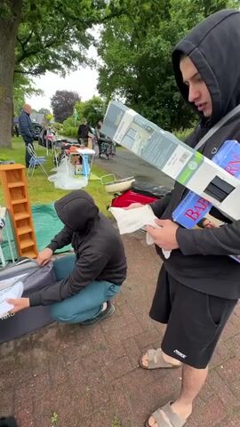 A point of view video shows a group of people at what appears to be an outdoor flea market or yard sale. In the foreground, a young man wearing a black hoodie and teal pants crouches over a gray "lionelo" branded bag, unpacking or packing items. To his left, a girl with braided hair, wearing a white long-sleeved shirt and a black vest, handles a pink handbag and some white fabric. Another young woman in a pink shirt stands behind her, smiling. In the background, several adults browse tables with various goods. One woman is looking at a brown wooden shelf. The ground is made of red bricks, and the area is surrounded by green grass and trees.