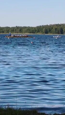 A long, narrow rowing boat with at least eight people on board, dressed in light-colored caps and tops, is actively rowing across a wide, blue body of water. The people are positioned in pairs, facing the stern of the boat, and are engaged in synchronized rowing. A coxswain stands at the stern, facing forward, presumably steering and coordinating the rowers.

The scene is set on a calm lake with gentle ripples on the surface, indicating a tranquil day. In the distance, a dense line of green trees forms the backdrop, suggesting a wooded area on the shore. The sky is a clear, pale blue, indicative of fair weather, likely during the daytime. Several orange buoys are visible in the water, marking a course or an area for the rowing activity. To the right of the main rowing boat, another, smaller boat with fewer occupants can be seen further in the distance, also appearing to be engaged in a water sport.

The overall impression is one of a sporting event or training session taking place in a natural, serene environment. The clear skies and calm water suggest a pleasant day for outdoor activities. While no text is visible in the image, the presence of multiple boats and buoys strongly suggests a competitive or organized event.