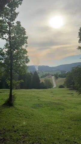 This media file captures a serene landscape view in Petit-Val, Switzerland. The scene is dominated by rolling green hills covered in lush grass, dotted with small wildflowers. In the foreground, a tall, leafy tree stands to the left, its shadow cast across the grassy slope.

In the middle ground, a winding road meanders through the valley, leading towards a cluster of buildings that appear to be a small village or farmsteads nestled among the trees. A plume of smoke is visible rising from the direction of the village, suggesting an ongoing activity such as a bonfire or perhaps a chimney. The distant hills are a patchwork of green forests and lighter-colored fields, with a few scattered houses.

The sky above is partially cloudy, with the sun shining brightly through a break in the clouds, creating a slightly hazy, warm glow. The sun appears high in the sky, indicating it is likely daytime, possibly late morning or early afternoon. The overall atmosphere is peaceful and idyllic, with a touch of rustic charm. There are no visible people or specific objects that draw significant attention, the focus being on the natural beauty of the Swiss countryside. The presence of the smoke is the only element suggesting human activity, subtly interacting with the otherwise tranquil environment.