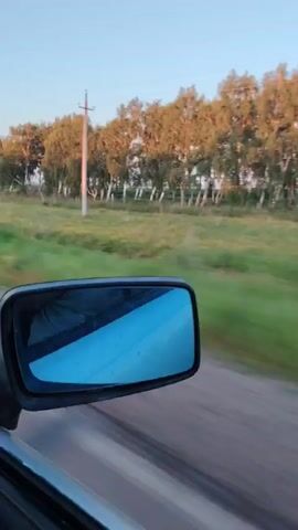 The video shows a side view mirror of a car. The mirror reflects the interior of the car, where a person wearing a black jacket is seated. The background shows a rural landscape with trees and fields. The sky is clear and blue. The car appears to be moving at a high speed, as indicated by the blur of the landscape in the background. The video is taken during the daytime.