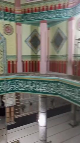 The video pans across the interior of a mosque undergoing renovation. Scaffolding is visible, along with renovation supplies. Decorative elements include Arabic calligraphy, geometric patterns, and red and green striped sections. The mihrab, or niche in the wall that indicates the direction of Mecca, is visible, along with a blue and white minbar, or pulpit. Light fixtures are also visible.