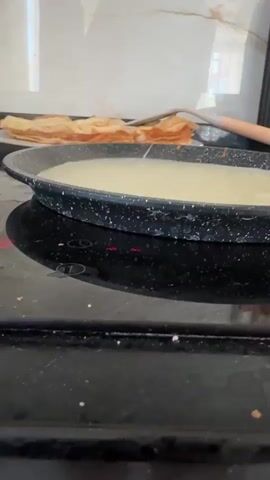 A close-up video shows a black frying pan on a black stovetop. The pan contains a light-colored liquid, possibly batter. In the background, a stack of golden-brown crepes or thin pancakes is piled on a white plate, with a wooden utensil resting beside them. Red indicator lights are visible on the stovetop beneath the pan, suggesting it is turned on and heating. The environment appears to be a kitchen.