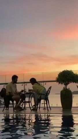 A man and a woman are seated at a small round table with latticework chairs on a terrace, engaged in conversation.  The man, sporting a beard and wearing a light-colored shirt and shorts, gestures as he speaks. The woman, clad in a light yellow top and patterned skirt, leans in, listening attentively.  A glass railing surrounds the terrace, and beyond it, a view of the ocean under a colorful sunset sky. In the foreground, the edge of a pool reflects the sky's hues. A potted plant stands to the right of the table.