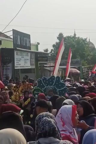The video captures a crowded street scene, likely a parade or festival. A large group of people, many wearing headscarves, are gathered, some holding Indonesian flags which are red and white. In the center of the frame, there's a decorated float with a green, floral-like design and a person sitting on it. To the left, another person is dressed in a costume with elaborate headdress. Buildings and trees are visible in the background, along with a distant domed structure, possibly a mosque or temple. The atmosphere appears festive and lively.