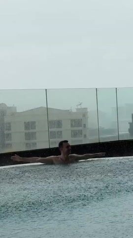 A man is in a swimming pool on a cloudy day. He is floating on his back with his arms spread out. Rain is falling in the pool. In the background, there is a glass barrier and a building.