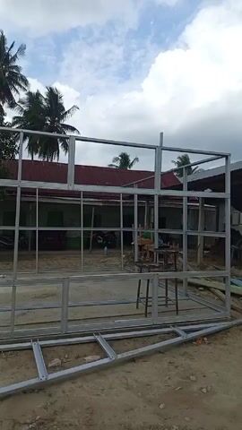 The camera pans to the left across the construction site of a building made with steel beams. The frame of the building is up but incomplete, with horizontal beams still on the ground. There are several motorcycles, tables, and chairs within the building, and what seems to be construction workers in the background. Several palm trees tower over the surrounding buildings, and the sky is cloudy.