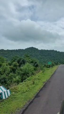 The video captures a landscape view from a vehicle. It shows a road on the right side, bordered by lush green grass and vegetation. A sign with green text on a white background and black and white striped pole is visible on the left side of the road. Beyond the road and grass, rolling green hills covered in trees dominate the background. The sky is overcast with white and grey clouds. The camera appears to be moving, as evidenced by the slight motion blur and the changing perspective of the landscape. There are no people or animals clearly visible in the frame. The overall impression is of a natural, possibly rural or forested, environment on a cloudy day.