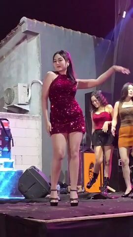 The video shows a group of women dancing on a stage. The woman in the foreground is wearing a sparkly red dress and high heels. She is dancing and smiling. Behind her, two other women are also dancing. The stage is lit with colorful lights. There are speakers and other equipment on the stage. The background appears to be an outdoor venue.