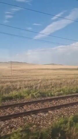 The video is shot from a moving train, showing a vast, golden field of dry grass stretching towards a gently rolling, barren landscape under a bright blue sky with scattered white clouds. Railroad tracks are visible in the foreground, indicating the train's movement. Power lines are strung across the sky, supported by poles. In the distance, a single tree stands in the field, and a solitary, dark hill or mountain range is discernible on the horizon. The lighting suggests it is daytime.