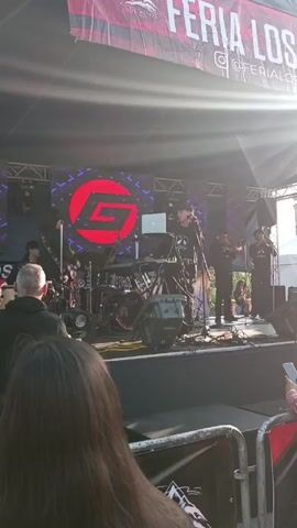 The video shows a live band performing on a stage at an outdoor event. The band consists of several musicians, including a singer, drummer, bass guitarist, and two horn players. The stage is set up with a large screen displaying a logo in the background. The event appears to be a festival or fair, with tents and people visible in the background. Several people are watching the performance, some of whom are recording with their phones. The atmosphere seems lively and festive. The bright sunlight suggests it is daytime. A banner overhead reads "FERIA LOS ALTOS". The barrier in the foreground is marked with "LOS ALTOS".