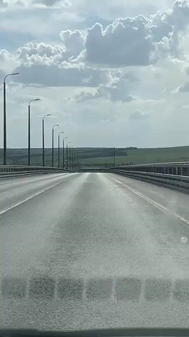 The video shows a car driving on a highway bridge during daytime. The camera is likely placed behind the car's windshield. The road is gray and made of asphalt. There are tall, gray lampposts on either side of the road. The sky is bright and light blue, with a few white, puffy clouds scattered across it. Beyond the edges of the bridge, the landscape looks flat and green with some vegetation. The overall impression is of a long, straight road stretching into the distance.