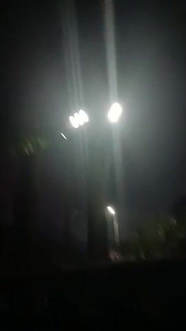This video appears to be captured at night, possibly in a rural or suburban area. The primary focus is a tall street light with multiple bright bulbs that illuminate the surroundings. The light creates strong beams that cut through what seems to be mist or light rain, giving the scene an ethereal quality. In the background, there are silhouetted shapes of houses and vegetation, suggesting a residential area. The lower portion of the video shows a dark foreground, possibly a railing or a fence, obscuring a clearer view of the immediate ground level. The camera movement is subtle, with some shakiness, indicating it might be handheld. The audio, if present, would likely consist of ambient night sounds, potentially including the gentle patter of rain or wind. The overall impression is one of a quiet, possibly damp, nighttime scene illuminated by artificial light.