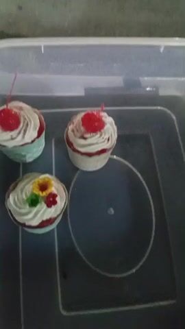 The video begins with a close-up of three decorative cupcakes in a clear plastic container. The cupcakes are frosted with white icing and topped with red cherries, except for one which has additional floral decorations. The container is placed on a dark, rough surface, possibly asphalt. As the video progresses, the camera pans up and to the right, revealing a blurry background of people and what appears to be a market or street vendor area at night. The lighting is dim, with some illuminated stalls in the background. The overall scene suggests an outdoor evening setting, possibly a food stall or market.