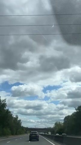 A first-person perspective video of driving on a highway under a cloudy sky. The camera is positioned behind a car and shows the road ahead, with other vehicles visible. A concrete overpass spans the highway in the distance. Trees line both sides of the road. The sky is filled with a mix of dark grey storm clouds and patches of blue sky with white cumulus clouds. There is some glare on the windshield.