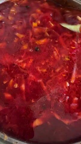This media file is a close-up shot of borscht, a traditional Eastern European soup, simmering in a pot. The soup is a deep, rich red color, indicative of the beet base, with visible chunks of vegetables such as cabbage, carrots, and possibly potatoes. A ladle is used to stir the soup, bringing some of the ingredients to the surface. The lighting suggests an indoor setting, possibly a kitchen, and there are no cues for time of day or weather. The focus is entirely on the food, with no people or explicit location markers other than the context provided. The activity is the cooking or preparation of the borscht. The prominent colors are shades of red and orange from the ingredients, against the darker broth. There is no visible text in the media file. The provided location context of Peremyshliany, Ukraine, suggests a cultural setting for this traditional dish.
