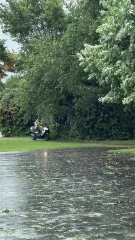 A person wearing a neon yellow reflective vest and helmet rides a silver scooter through a large puddle on a paved area. The area is surrounded by green trees and grass. The day is overcast and rainy.