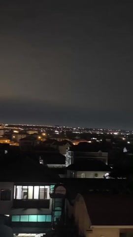 This is a video of a cityscape at night, likely from an elevated vantage point. The foreground shows the rooftops of several houses, with some windows illuminated by interior lights. Beyond these houses, the city stretches out into the distance, a vast expanse of scattered lights against the dark sky.

The time of day is clearly night, with the sky a deep, uniform gray, indicating cloud cover or a lack of moonlight. The lights of the city suggest human habitation and activity. In the far distance, a tall tower, possibly a radio or transmission tower, is visible, illuminated with red lights near its top. There are also other structures and clusters of lights that hint at the urban landscape.

The setting appears to be a residential area bordering a larger city. The houses in the foreground seem to be detached or semi-detached, with pitched roofs typical of many residential structures. The overall mood is calm and somewhat quiet, as is common in a city at night. There are no people or specific events visible. The focus is on the expanse of the city and the interplay of artificial light against the natural darkness.

There is no visible text in this video.