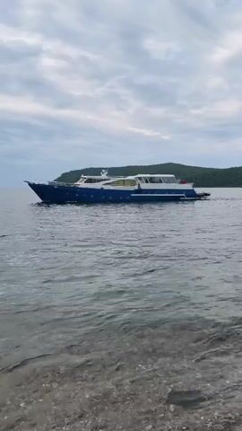 A blue yacht sails on calm water under a cloudy sky. The foreground shows the rocky shore of a body of water. The yacht is seen from the side, moving from left to right across the frame. The background features a green, tree-covered landmass on the horizon. The lighting suggests it is either early morning or late evening.