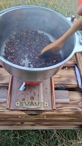 The video shows a close-up shot of coffee beans being roasted in a metal pot over a burner. The pot is placed on a wooden stand with copper accents and a brand name "EVAVE" inscribed on it. A person's hand is visible, holding a wooden spoon and stirring the coffee beans. The beans are dark brown and appear to be at various stages of roasting, with some steam rising from the pot. The background suggests an outdoor setting with grass and water, possibly a lake or river, visible in the distance. The overall lighting indicates it is daytime.