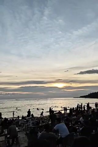 The video shows a beach at sunset. Many people are either sitting at tables on the sand or swimming and wading in the ocean. The sun is setting on the horizon, visible behind clouds. The sky has a blue and gray hue. A sailboat appears in the water.