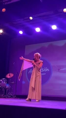 A woman in a long, light-colored dress and a scarf stands center stage on a platform, holding a microphone. She gestures with her left arm, her scarf trailing. Behind her, a large screen displays a circular logo that reads "AulaVisión Song Contest" with images of hands and clouds. To the left of the stage, a drum set is visible. The stage is lit with purple spotlights. The woman is the sole focus of the video, and she is the only person visible on stage.