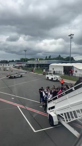 The image captures an airport tarmac scene in Tillé, France, under an overcast sky with heavy gray clouds. A group of approximately 15-20 passengers is in the process of boarding an aircraft (which is not fully visible) via a mobile set of air stairs. Some individuals are already ascending the white metal stairs, while others are queued at the base, waiting their turn.

In the background, an airport terminal building with multiple windows and a flat roof is visible. Several airport vehicles are parked on the tarmac: a flatbed baggage or cargo trolley to the left, and a white utility van or car further back. An airport staff member, wearing an orange high-visibility vest, stands near the white van. Tall light poles with multiple lamps are positioned across the tarmac. The ground features various white and red markings, typical of an airport apron. The overall impression is a routine boarding operation during daytime hours at a regional airport.