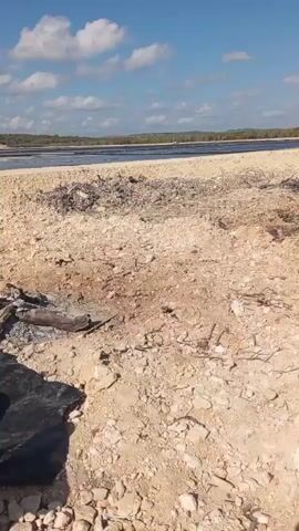 The video shows a group of people sitting on the ground in an outdoor environment near a body of water. The ground is sandy and rocky, with scattered debris, including what appears to be remnants of a campfire. Some individuals are wearing work clothes, including hats and vests. The body of water in the background is calm and appears to be a salt flat or shallow lake. The sky is blue with scattered clouds.
