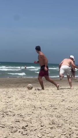 The video shows two men playing soccer on a beach on a sunny day. The man closest to the camera is wearing dark swim trunks with a red and black pattern. The other man is wearing white shorts and a white baseball cap. They are both shirtless. The beach is sandy, and the ocean waves are visible in the background. A boat on wheels passes them.