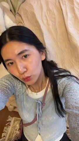 A young Asian woman is lying on a tan bedspread. She is wearing a light blue lacy shirt over a white top, with a pink purse strap visible on her shoulder. Her dark hair is pulled back, and she is wearing a necklace. She puckers her lips and winks at the camera. A blurry white dog statue sits on a dark wooden table behind her.