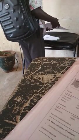 A person with braided blonde hair wearing a white floral shirt and dark pants is seated at a table, holding and examining a pink document. A black backpack is visible on the chair behind the person. The environment appears to be indoors, with a wall in the background and a potted plant on the floor in the foreground. The document has printed text and a photograph.