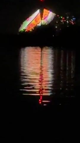 The video shows a nighttime view of a body of water with a colorful, glowing structure reflected on the surface. The structure appears triangular or winged, with illuminated stripes and possibly some additional lights. The water reflects the light in a shimmering pattern. The surrounding area is dark. The overall scene is low-light and somewhat grainy.