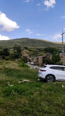 A video shot from a low angle shows a rural landscape. In the foreground, a white car is parked on the side of the road, partially obscured by tall grass. The car is a modern SUV with a sleek design. Behind the car, a few other vehicles are visible further down the road. The road leads to a village of stone houses with rustic architecture. The houses appear to be old, with some showing signs of wear and tear. In the background, rolling hills covered in green vegetation rise up towards a clear blue sky with a few scattered clouds. The overall impression is one of a quiet, peaceful village nestled in a beautiful natural setting.