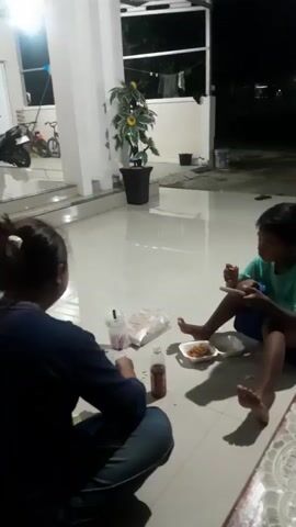 This is a video showing two people, likely a child and an adult, sitting on the floor of a porch or patio area. The floor is tiled and appears to be wet, suggesting it may have recently rained or been cleaned. A potted plant with large, yellow flowers stands against a white wall. In the background, there are glimpses of a bicycle and a motorbike, indicating a residential setting.

The child, wearing a green t-shirt and blue shorts, is seated on the floor with their legs extended. They are looking down at a smartphone in their hands. A plate of food, appearing to be fried items, is placed in front of them. Next to the plate are several small white packets, possibly containing snacks or condiments.

The adult, seen from behind, is wearing a dark blue top and jeans. They are also sitting on the floor, facing away from the camera. In front of them are a plastic cup with a straw, a bottle of what appears to be a beverage, and other items that are difficult to discern due to the low lighting and motion blur.

The scene is dimly lit, suggesting it is either evening or nighttime. The overall atmosphere appears casual and relaxed, with the child engrossed in their phone while the adult seems to be preparing or consuming food and drink. The location cues, such as the architectural style and the presence of common household items, are consistent with a setting in Panjalin Kidul, Indonesia. There is no visible text in the video.