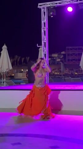 A woman performs a belly dance on a stage at night. She is wearing a sequined orange bra, a matching flowing skirt, and jewelry on her wrists. The stage is lit with colorful lights that change between white, blue, purple, and yellow. Behind the stage are resort amenities, including a pool, umbrellas, and water slides. The dancer smiles and moves gracefully as she follows the music.