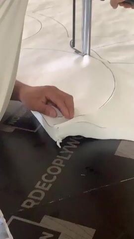 The video shows a close-up view of a person cutting a large piece of white material with a hot knife. The material is laid out on a dark surface, possibly plywood, with markings or lines drawn on it. The person's hand is visible, holding down the material near the cutting line to ensure a clean cut. The hot knife is creating a clean, melted edge as it moves along the traced pattern. The scene appears to be a workshop or manufacturing environment where custom shapes are being cut from the material. The overall impression is one of precision and focused work.