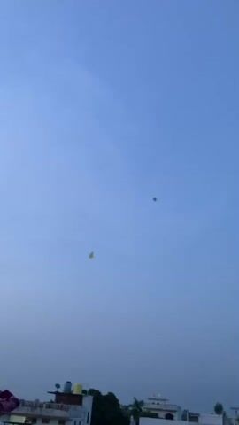 A video shows a group of people on a rooftop, flying kites against a hazy blue sky. In the foreground, a person with dark hair, wearing a pink and white patterned shirt, looks up at the sky. To the left, a yellow kite is visible in the air. Further to the right, a green kite is also flying. In the mid-ground, buildings and trees are visible. As the video progresses, more people appear on the rooftop, including two men and another person. One man, wearing a white shirt, is seen talking to another man in a green shirt, who is facing away from the camera. The sky displays a gradient of blue, with a hint of pink near the horizon in the later frames. The overall scene depicts a casual gathering of people enjoying kite flying on a rooftop.