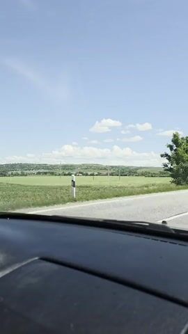 The video shows a first-person perspective from inside a moving vehicle, looking out the front windshield. The car is driving on a paved road with a white dashed line on the right side. On the right side of the road, there are green trees and a white and black striped pole visible. Beyond the road, on both sides, are vast green fields under a clear blue sky with some scattered white clouds. In the distance, hills and a small town with several buildings can be seen.