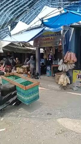 A busy outdoor market scene. The camera is positioned on the right side, focusing on a motorbike with a large green woven basket attached to the rear. In the foreground, the ground is covered with gravel and dirt. To the left of the motorbike, another green woven basket is visible. The market stalls are covered by a blue tarp and a black mesh tarp, creating shade. Several people are seen in the background, some appearing to be vendors and others customers. One stall displays various packaged goods, including what appear to be snacks or spices, with a sign reading "PENGGILIGAN SAMBAL KACANG". Another stall has a selection of fruits or vegetables in woven baskets. A yellow sign with red text and an image of a smiling face is also visible. A motorcycle license plate reads "K 3132 QJ". The overall impression is one of a vibrant, bustling local marketplace on a sunny day.