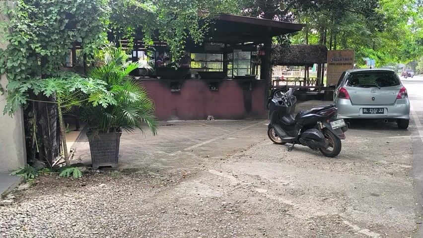 A parked silver car and a motorcycle lying on its side are on a concrete area. The motorcycle is parked close to the car. The other side of the concrete area shows a building and greenery. The building's facade appears dark and possibly charred. There are a number of tables inside the building. Various green plants are visible in the foreground. The video is shot in landscape mode. The overall impression is of a vehicle parking area and possible fire damage.