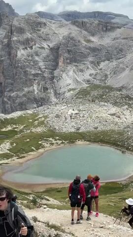 The video shows a panoramic view of a mountainous landscape with a turquoise lake in the foreground. Several hikers are visible, some standing and others sitting on the ground. The mountains are steep and rocky, with some vegetation on the lower slopes. The sky is partly cloudy, with patches of blue visible. The camera pans across the scene, capturing the grandeur of the natural surroundings and the presence of the hikers.