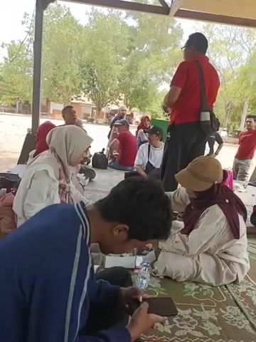 The video shows a group of people sitting on mats under a shelter. A man in a red shirt is standing and talking to a group of people. Some people are eating and drinking. There are trees and buildings in the background.