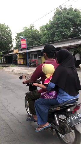 A man wearing a maroon shirt and a black baseball cap is driving a motorcycle on an asphalt road. He is wearing dark pants and flip-flops. Seated behind him is a woman wearing a black hijab and a blue shirt. Between them sits a young child wearing a yellow headscarf and a purple and blue patterned shirt. The child's legs are extended and bent at the knees. The motorcycle has a license plate that reads W 5136 NT. The background shows a row of buildings with awnings, some with signs, and trees. The sky is overcast.