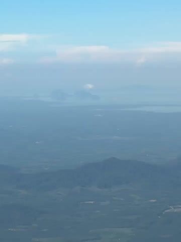 An aerial view shows a landscape with mountains and bodies of water under a hazy sky. In the foreground, rolling hills covered in dark green vegetation are visible. Several smaller, rounded hills are scattered throughout this area, some with hints of cleared patches that could be farmland or structures. A winding road can be faintly seen in the middle distance. Further back, a large, hazy body of water stretches across the horizon, dotted with several islands or landmasses of varying sizes. The sky above is a pale blue, with thin, wispy clouds that contribute to the overall muted and atmospheric quality of the scene. The perspective suggests the video was filmed from a high altitude, possibly from an aircraft.