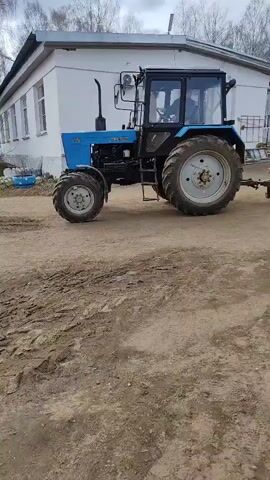 Here's a description of the video:

The video shows a light blue tractor parked on a dirt area next to a white, one-story building. The tractor has the word "Беларус" written on its side, with the model number "521" below. The tractor is pulling a small trailer. A person is visible inside the tractor's cab. The ground is muddy. In the background are trees and a cloudy sky.