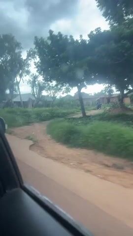The video shows a view from the inside of a car looking out the passenger side window. The car is driving on a dirt road. On the left side of the road there is a grassy area with some trees. On the right side of the road there are a few buildings, some motorcycles and people standing around. The sky is cloudy.
