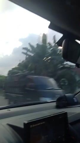 The video is filmed from inside a vehicle, looking forward. The time appears to be late afternoon or early evening as the sun is low in the sky, casting a warm glow. The road ahead is visible, with a curve to the left. On the left side of the road, there are houses and trees. A person is riding a motorcycle on the left side of the road, moving away from the camera. On the right side of the road, there are more trees and some structures that appear to be small shops or stalls. The sky is partly cloudy. The interior of the car is also visible, including the dashboard and a rearview mirror. The windshield of the car has some glare from the sun.