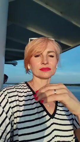A woman with short, peachy-pink hair and red lipstick poses for a selfie. She is wearing a black and white striped v-neck shirt and a necklace with a small pendant. Her right hand is touching her chin and neck. She is outdoors with a body of water and trees in the background, suggesting she might be on a boat or a waterfront. The lighting suggests it is either sunrise or sunset.