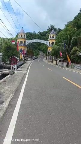 The video shows a road leading up to an archway with two yellow towers on either side. The archway has text on it, but it is difficult to read. There are Indonesian flags and other decorations on the sides of the road. There are some trees and buildings on either side of the road. A motorcycle is seen riding up the road. The sky is cloudy. The video is filmed from a moving vehicle.