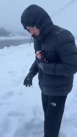 A young man, wearing a black puffer jacket, black sweatpants, and gloves, stands in the snow next to a road. He digs into the snow and retrieves a six-pack of beer, partially buried. He holds it up, smiling, then turns and walks toward a parked car. It is foggy and snowy.